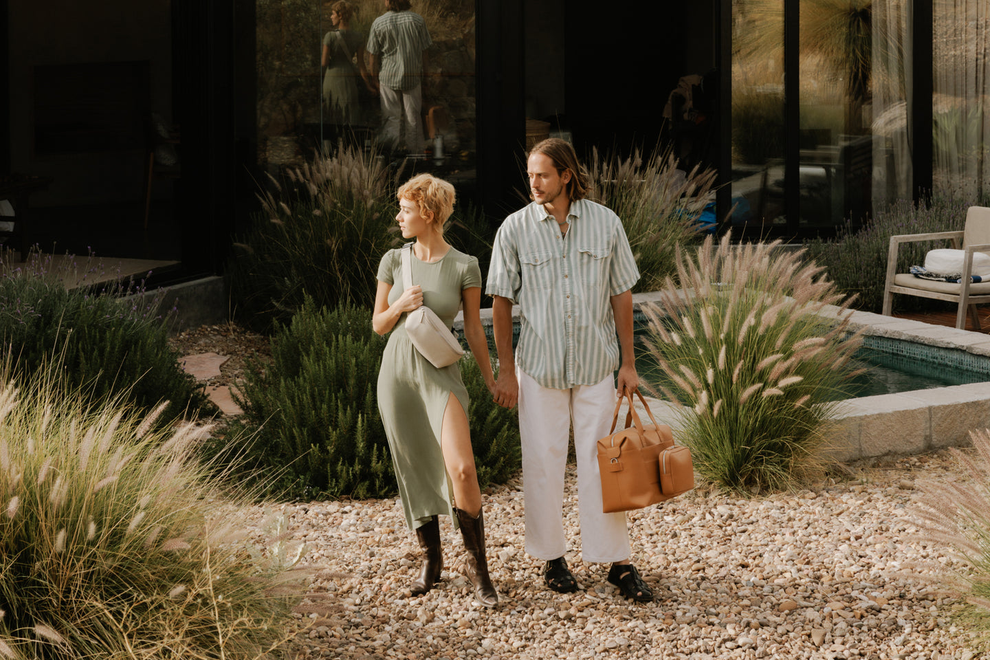 A stylish couple stands hand-in-hand outside, surrounded by tall grass and greenery. The woman is holding a Metro sling in ivory (vegan leather) while the man carries a Metro duffel in saddle tan (vegan leather). Both are dressed in casual yet elegant outfits.
