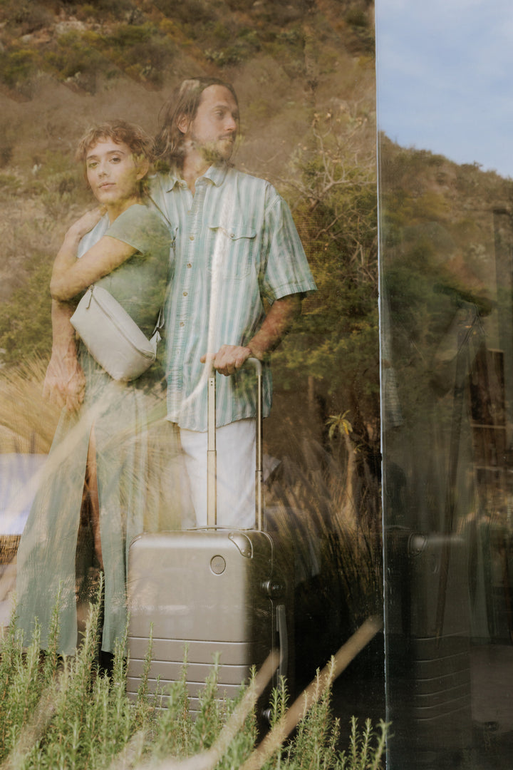 A reflective glass image of a couple standing close together, with the woman wearing a metro sling in ivory (vegan leather) and the man with his hand on a hybrid carry-on in champagne. Behind them, lush outdoor scenery is visible through the glass.