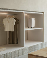 Close-up of an open shelving unit displaying a carry-on in desert taupe on the top shelf, a sevilla top in cream and sevilla dress in moss on hangers. The shelves are built into the wall, with a minimalist design.
