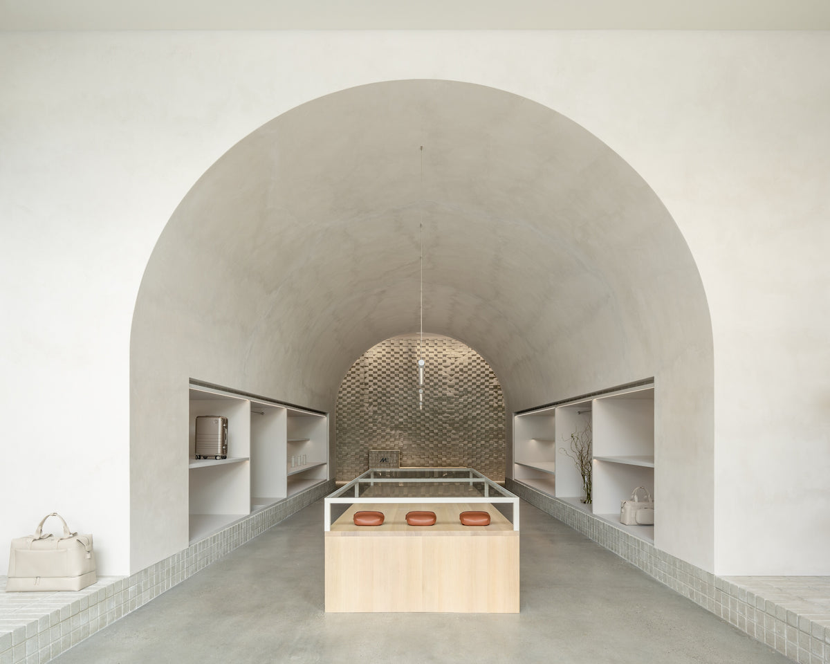A wide view of Monos retail store with a large archway ceiling, built-in shelving along the walls, and a central display table. The shelves contain a hybrid carry-on luggage, metro bags, and decorative items, with a tiled wall feature in the background.