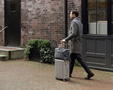 A man dressed in a grey coat walking on a brick pathway while pulling a Hybrid carry-on in silver with a metro duffel in dover grey secured on top.