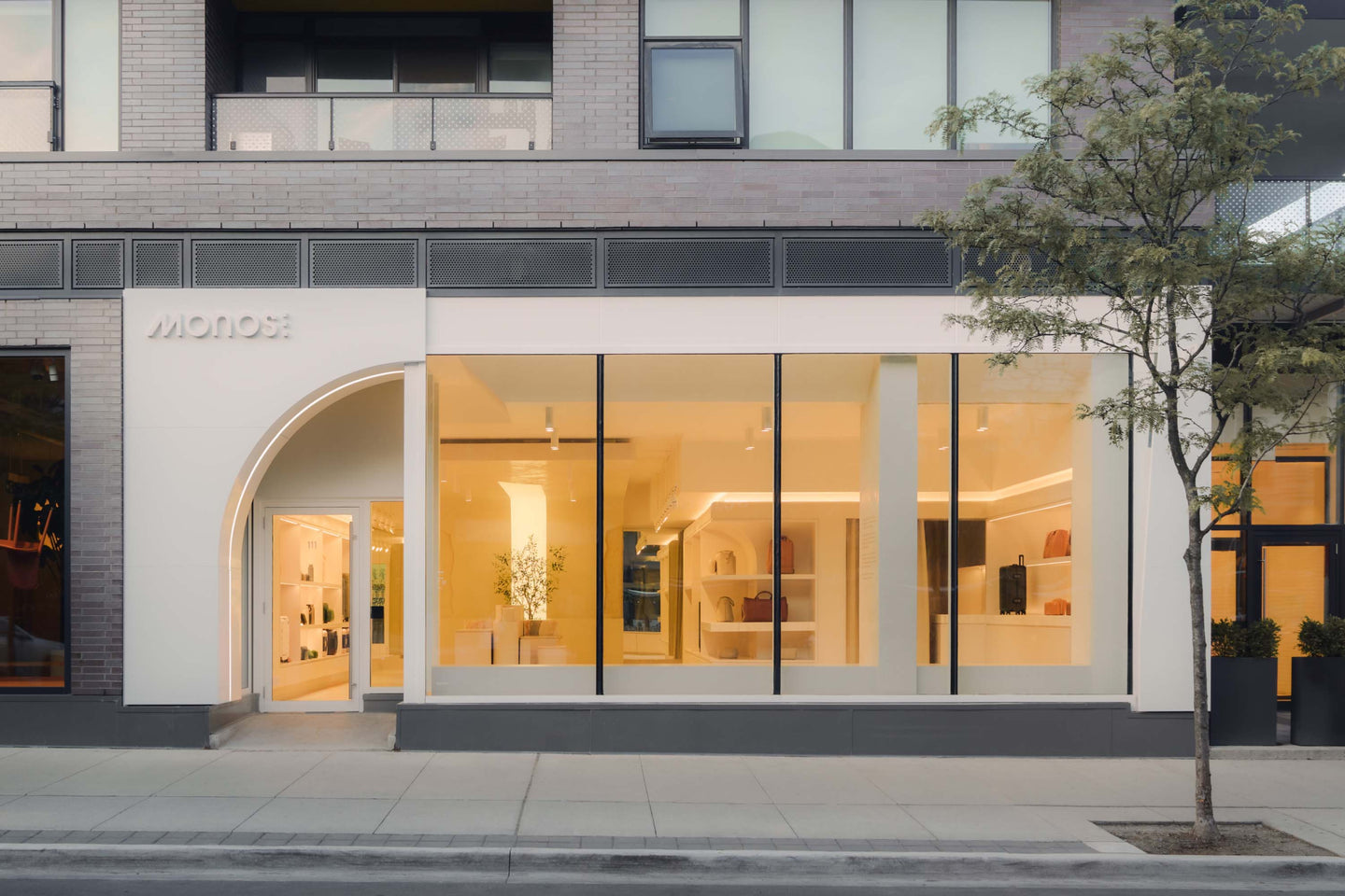 The exterior of the Monos Ossington retail store, featuring large windows that showcase a bright and modern interior with minimalist displays. A tree is visible on the sidewalk outside the store.