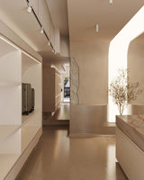 A softly lit, minimalist showroom with smooth, beige-toned walls. On the left, white shelves display a carry-on pro and check-in medium luggage, while the right side features a small tree planted in a recessed alcove, illuminated by natural light.