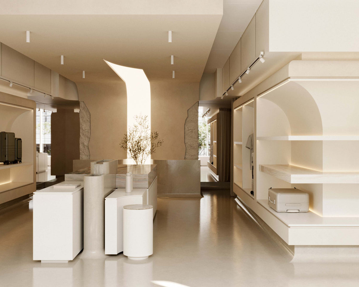 A stylish, open-concept showroom with neutral tones, featuring a central display table, Monos suitcases on shelves, and a softly lit tree in the background.