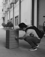 A man squatting and inspecting the packaging of a Monos Carry-On box, outside a factory building.