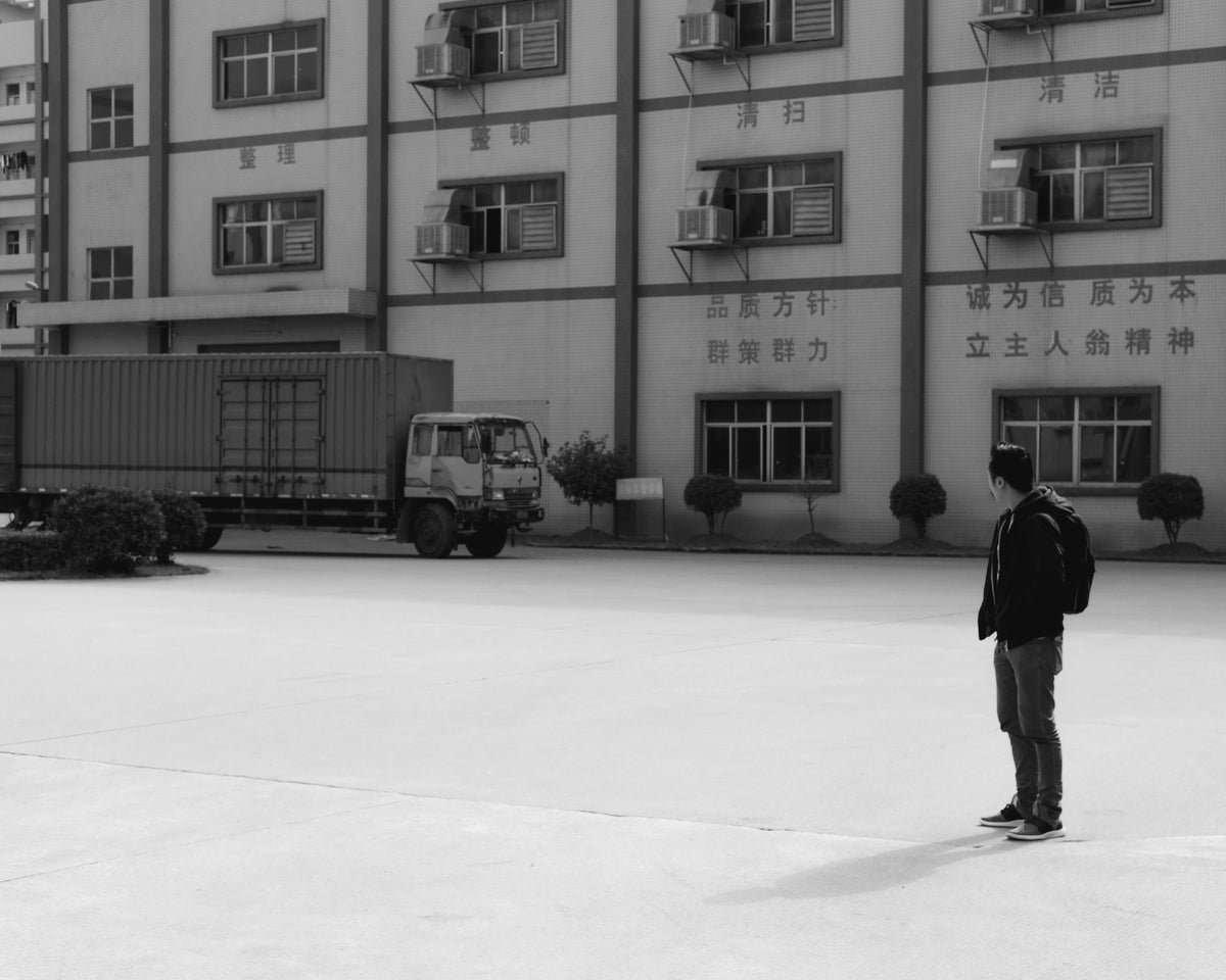 A man standing outside a large industrial building, with a logistics truck parked nearby.