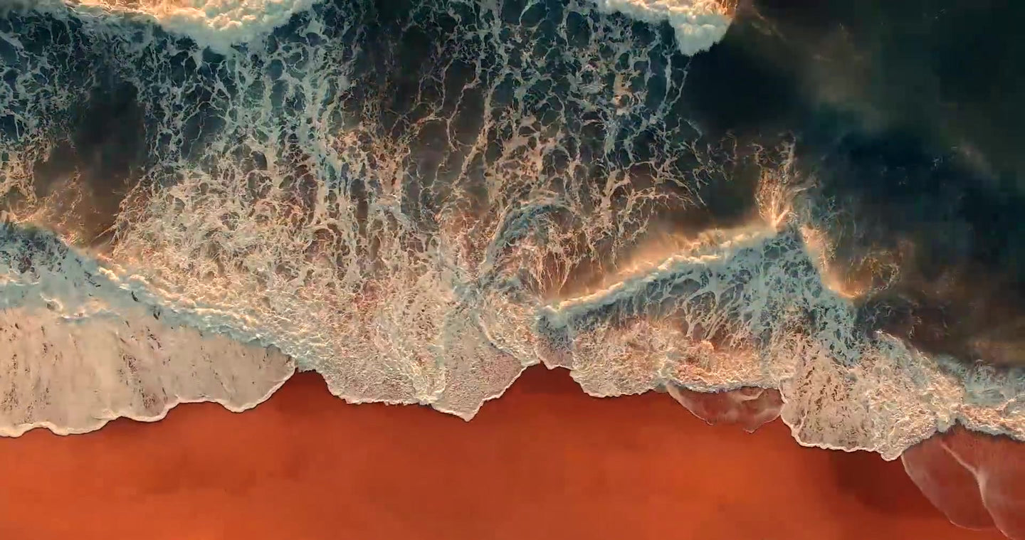 Aerial view of ocean waves crashing onto a sandy beach, with a gradient of golden light reflecting on the water. The waves create white foam as they meet the shoreline, blending into the deep blue sea.