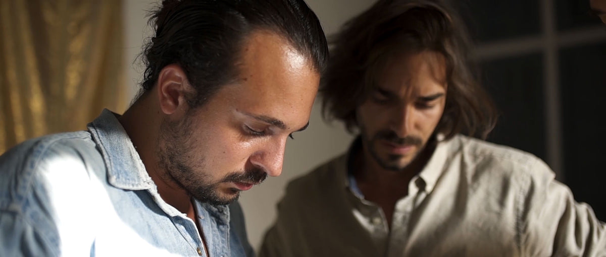 Two men in close-up focus. The man on the left is looking down thoughtfully, while the man in the background on the right is slightly out of focus, also looking down with a serious expression. The image suggests a moment of concentration or discussion.