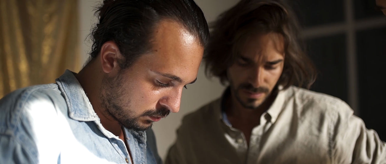 Two men in close-up focus. The man on the left is looking down thoughtfully, while the man in the background on the right is slightly out of focus, also looking down with a serious expression. The image suggests a moment of concentration or discussion.