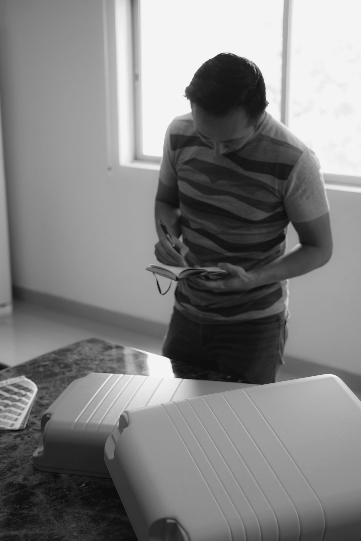 Black-and-white image of a man standing at a table, focused on writing notes in a small notebook. Two Monos suitcase shells are on the table in front of him, and a window in the background provides natural light. The scene suggests a design or quality inspection process.