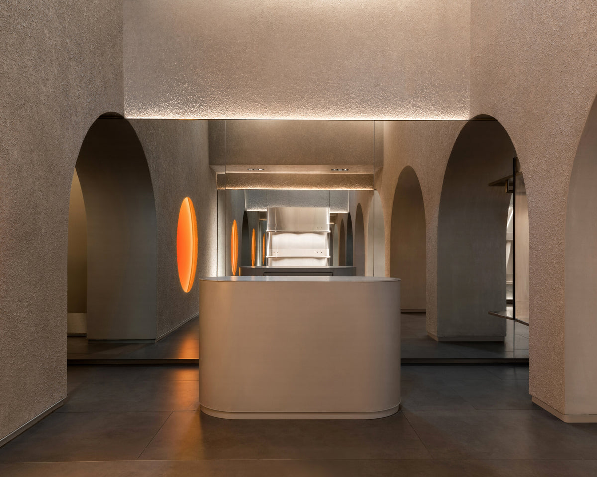 The reception area of the Monos Georgetown store, framed by textured stone arches and softly glowing circular light features. A curved minimalist counter stands at the center, reflecting the warm tones of the surrounding mirrors and brushed steel surfaces.