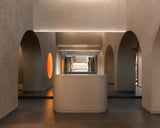 The reception area of the Monos Georgetown store, framed by textured stone arches and softly glowing circular light features. A curved minimalist counter stands at the center, reflecting the warm tones of the surrounding mirrors and brushed steel surfaces.
