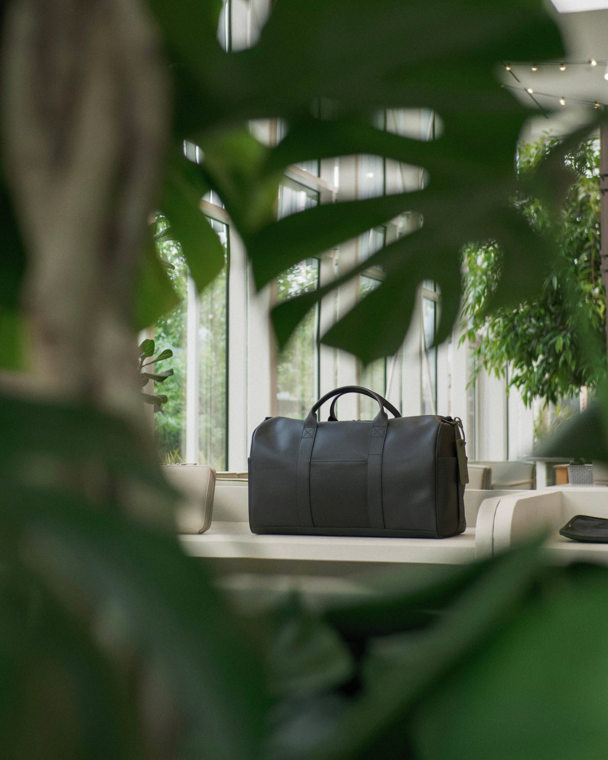 The Metro Carry-All Duffel in Carbon Black sits on a white bench in a modern interior, framed by large green plant leaves in the foreground.