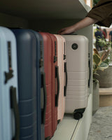 The Classic collection displayed on a shelf with a person's hand resting on a Desert Taupe suitcase.