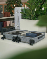 A Carry-On in Terrazzo Graphite is shown open on a white table, alongside a closed matching suitcase and other luggage pieces in a styled indoor setting with plants.