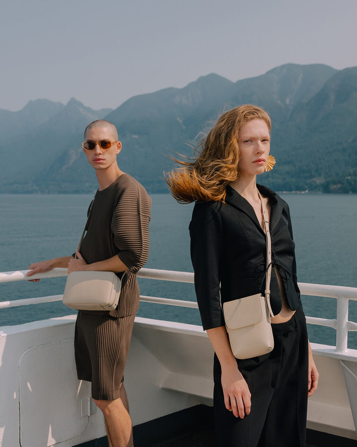 Two models on a boat deck with mountains and water in the background, wearing the Metro Mini Crossbody and Metro Crossbody in Ivory.