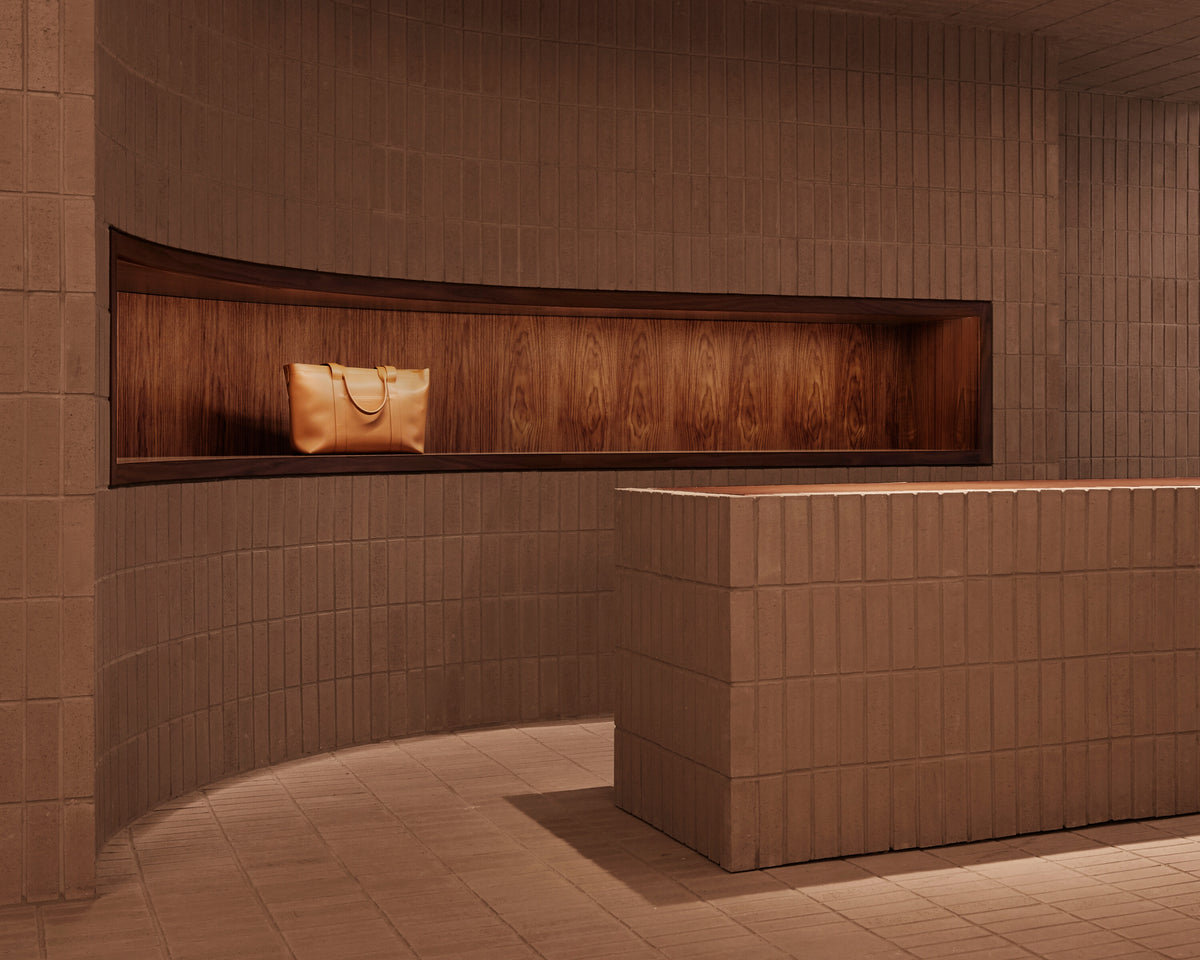  The Metro Tote in Saddle Tan displayed within a recessed wood alcove inside the Monos SoHo store, framed by curved terracotta brick walls and warm ambient lighting.