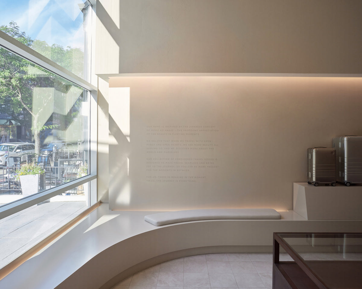 Monos Boston store interior with curved seating by a large front window and silver luggage displayed against a minimalist wall.