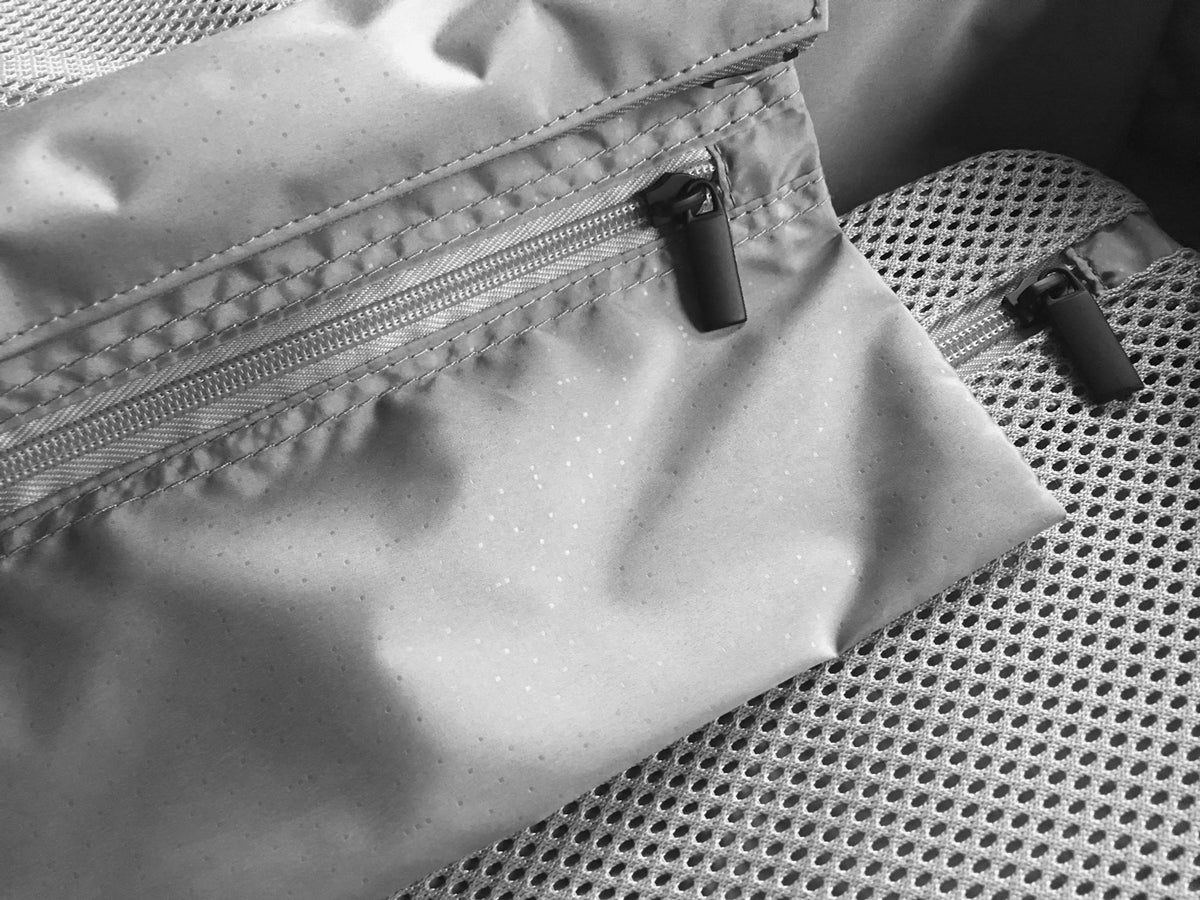 A close-up black-and-white image shows the detail of a zippered pocket on a Monos suitcase interior, with a smooth fabric texture and a sturdy zipper pull. The mesh lining is visible in the background.